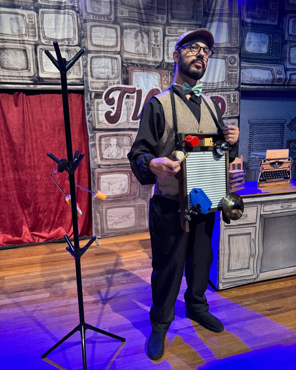 Ator de colete e gravata-borboleta segura uma tábua de lavar roupa cheia de instrumentos de percussão, diante da parede de TVs e de uma cortina vermelha